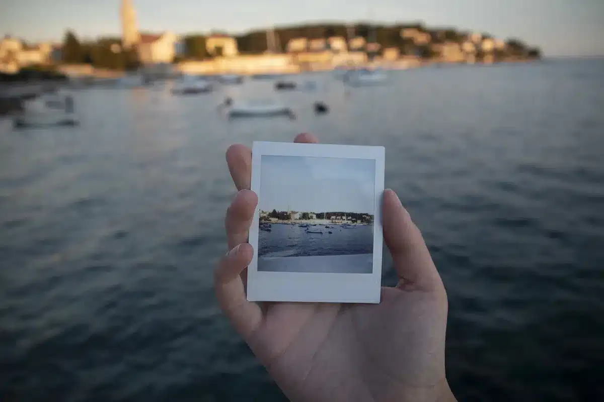 Hands holding a worn paperback and a Polaroid on a café table, people blurred in background